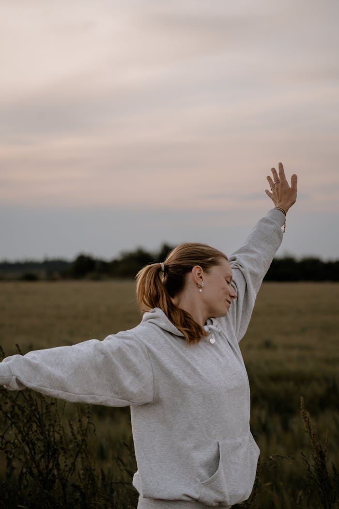 A woman enjoys nature, arms raised, in a peaceful countryside setting at dusk.